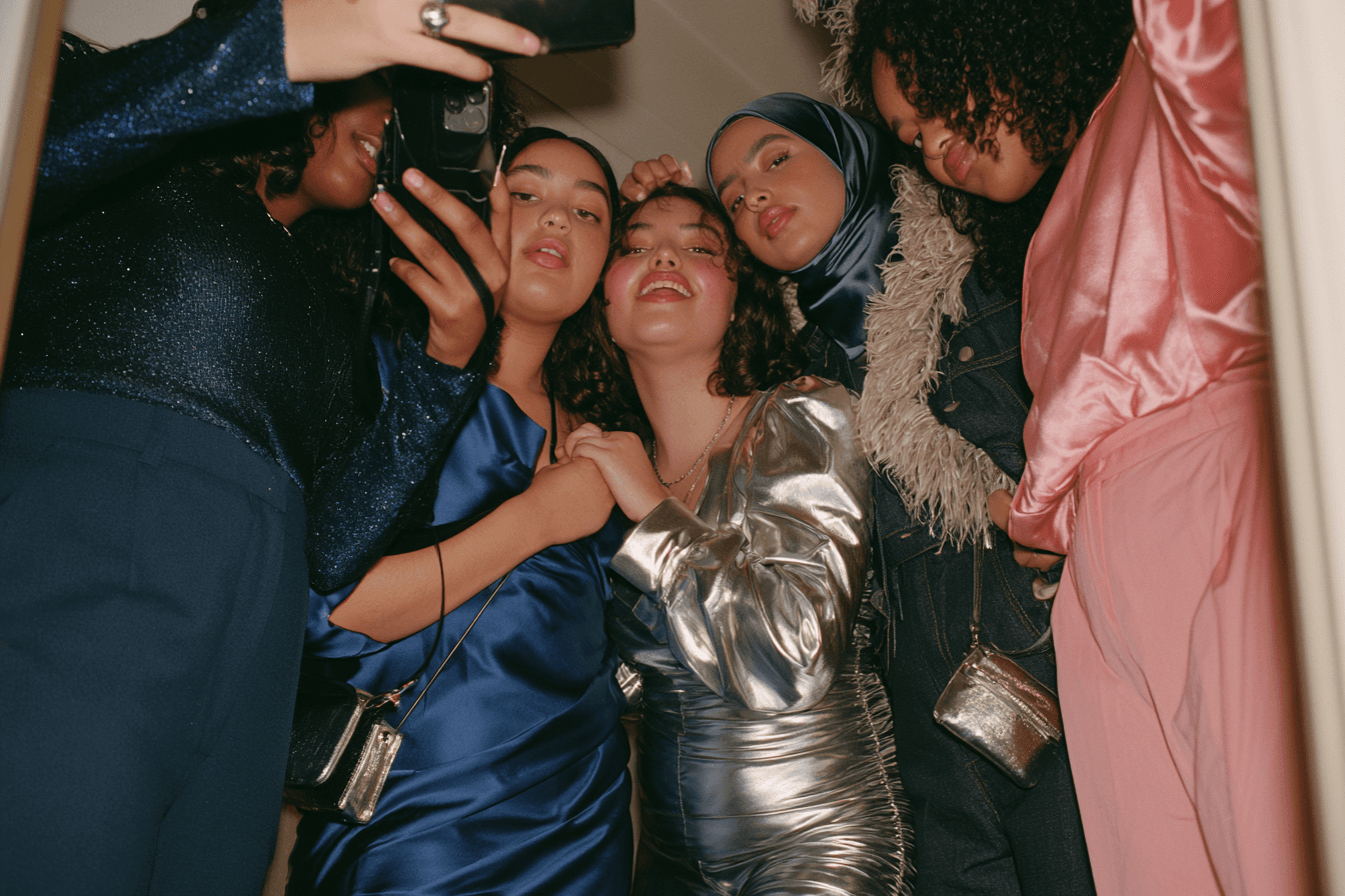 Seven friends crammed around a restaurant table and its mirror behind, phones up, sequins and satin and one feather trim, cobalt and hot pink and chrome across the group. Someone laughing mid-frame, someone fixing a strap. UK 8 to 26 visible in one frame, at least two in trousers or a jumpsuit, one hijab-wearing woman, one person with a mobility aid. Direct flash, unposed, genuinely chaotic.